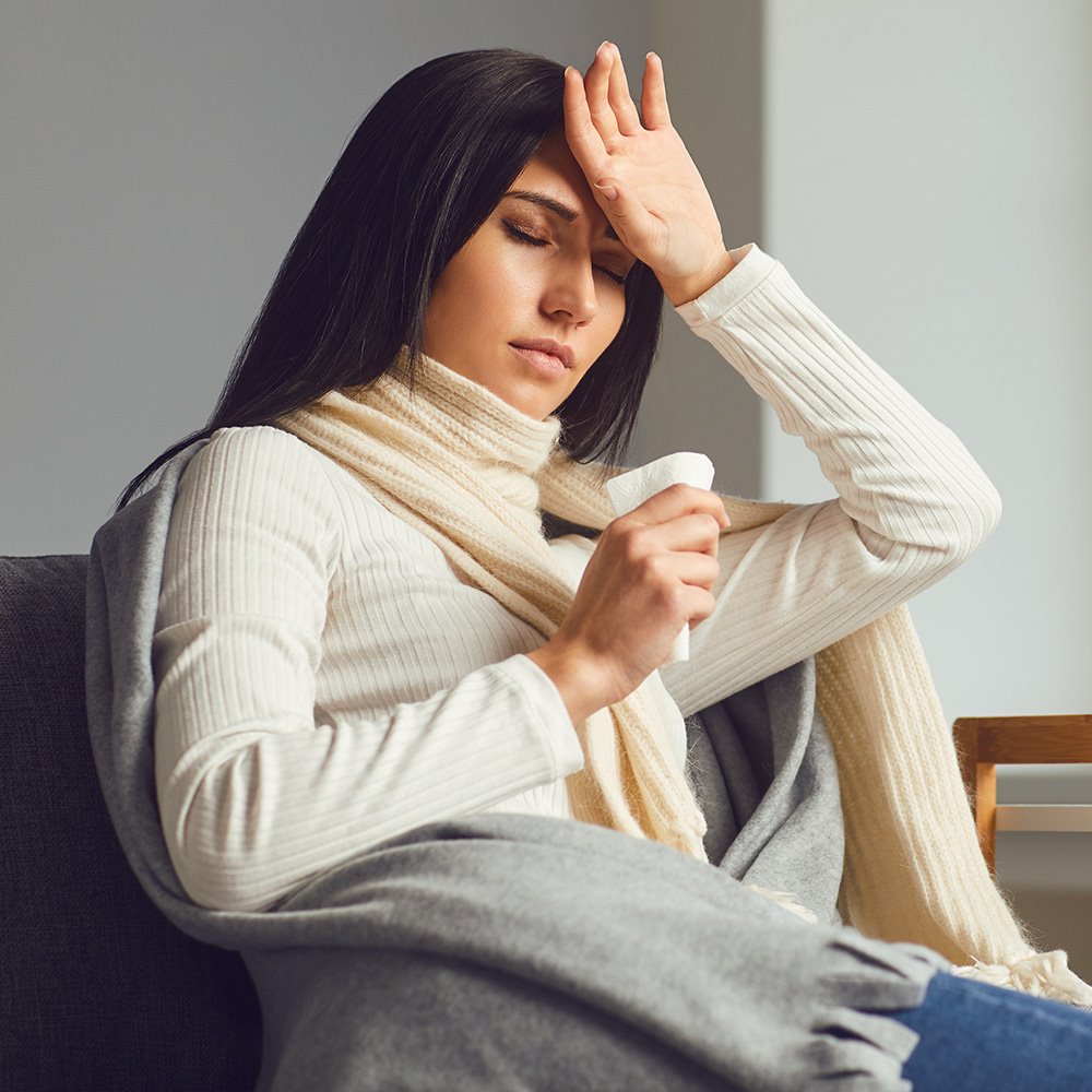 Woman who is feeling under the weather checking her temperature with the back of her hand and a tissue in the other hand while sitting on a couch at home