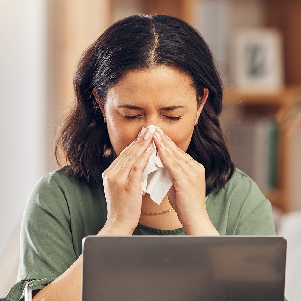 Women with allergies sitting in front of a laptop while blowing her nose