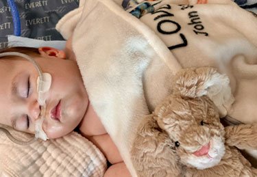 Baby sleeping while wearing a nasal cannula, covered by a blanket with a stuffed animal set next to the baby