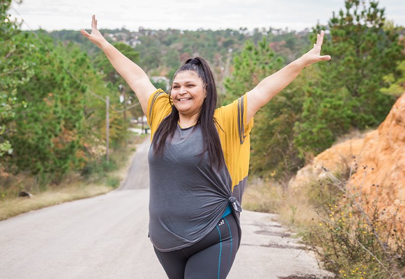 A smiling ARC patient stretching her arms wide during a walk along an Austin road, representing wellness and an active lifestyle