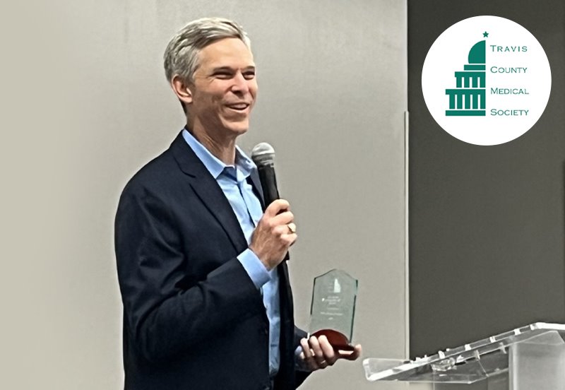 Dr. Mark Ambler giving an acceptance speech after receiving the TCMS 2025 Humanitarian of the Year award and the Travis County Medical Society logo