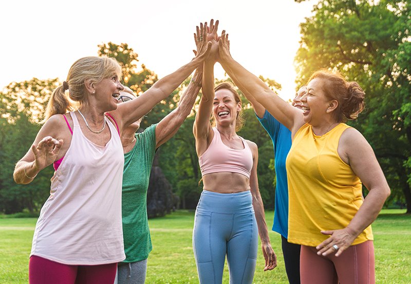 A group of women high-fiving outside at a park after exercising, representing an active lifestyle, preventive care, and Breast Cancer Awareness Month