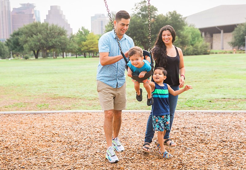 Mom, dad, and their two sons having a good time at the park while dad pushes his son on the swing, representing good health
